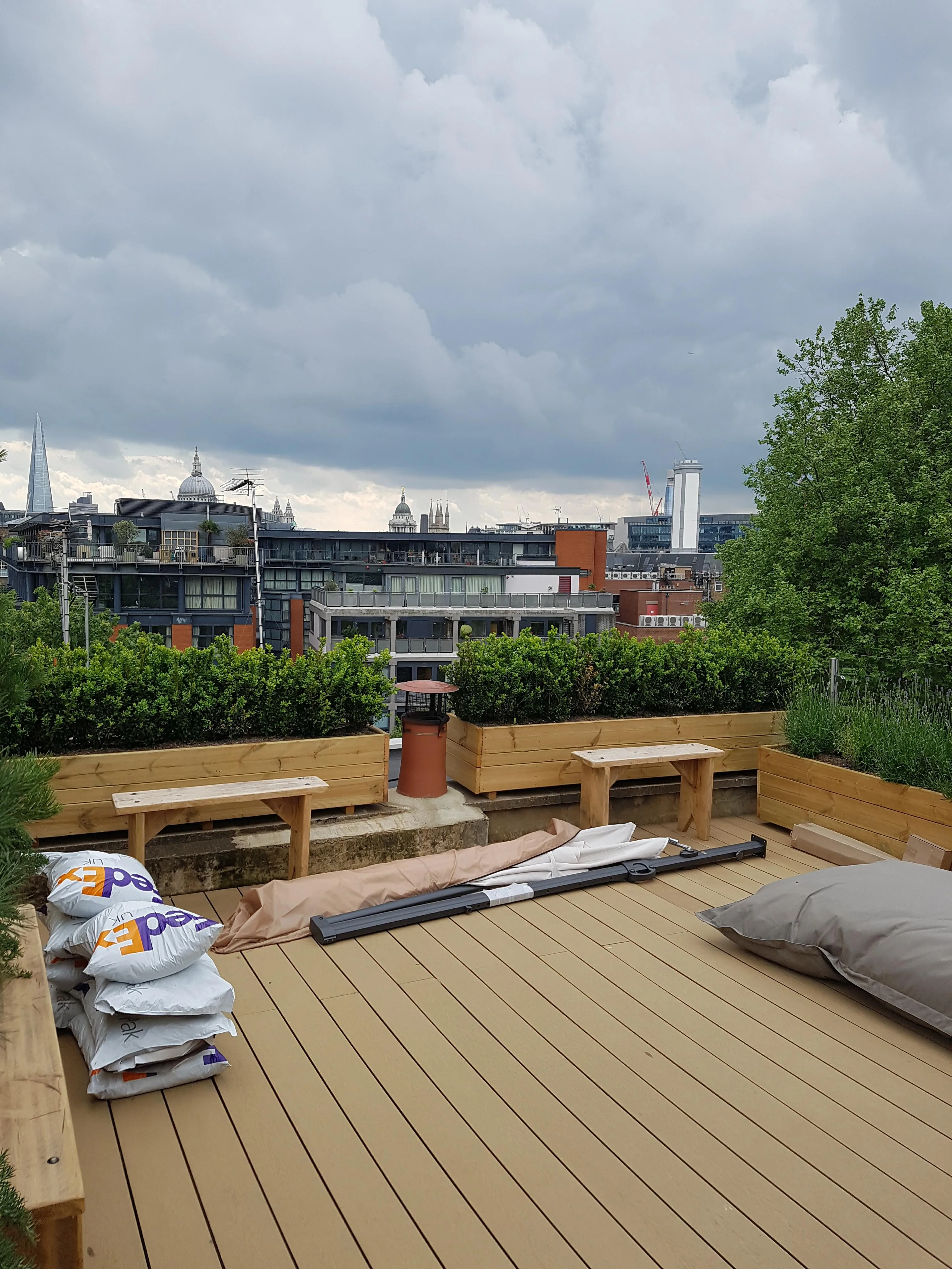 A rooftop patio featuring wooden decking and planters with greenery, overlooking a city skyline